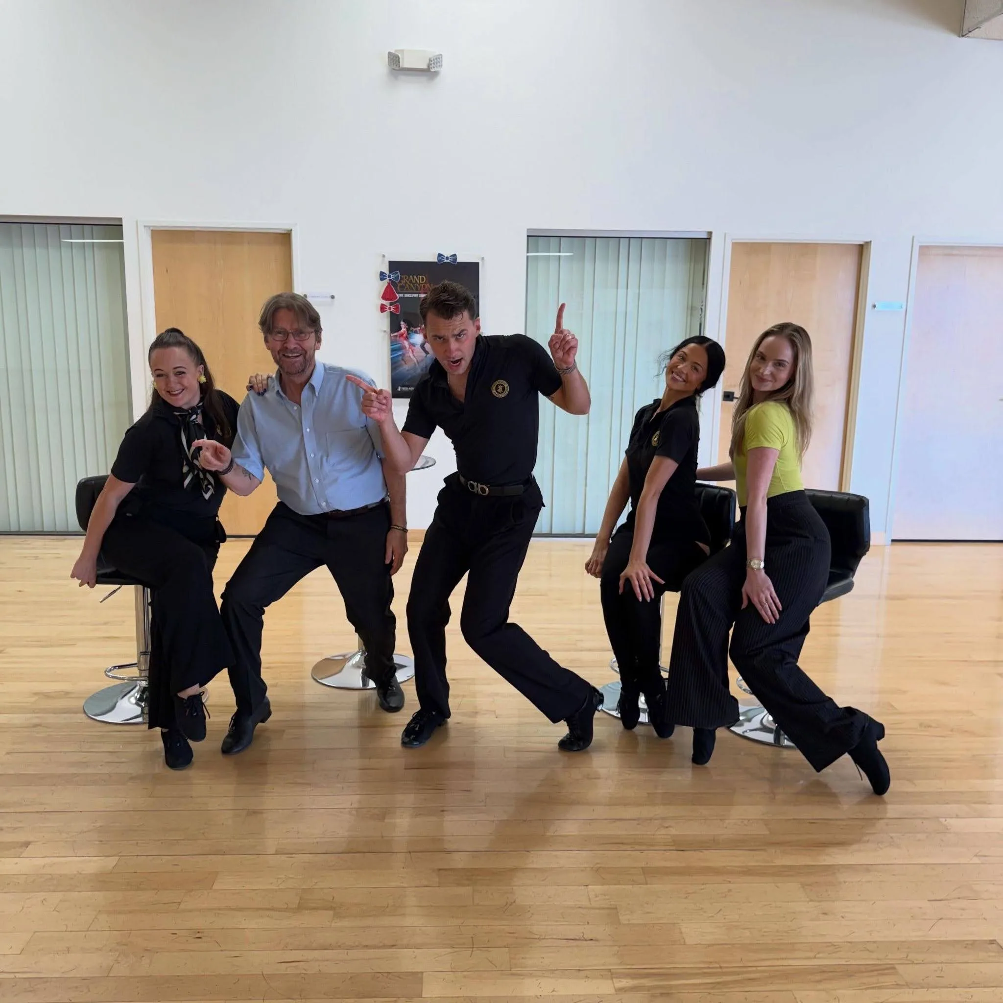 Five smiling people pose playfully inside a bright ballroom dance studio, showcasing a mix of comfortable, stylish outfits suitable for dance classes, from casual tops and slacks to dance shoes. A fun group photo illustrating what to wear to dance lessons in Ahwatukee for comfort, movement, and confidence.