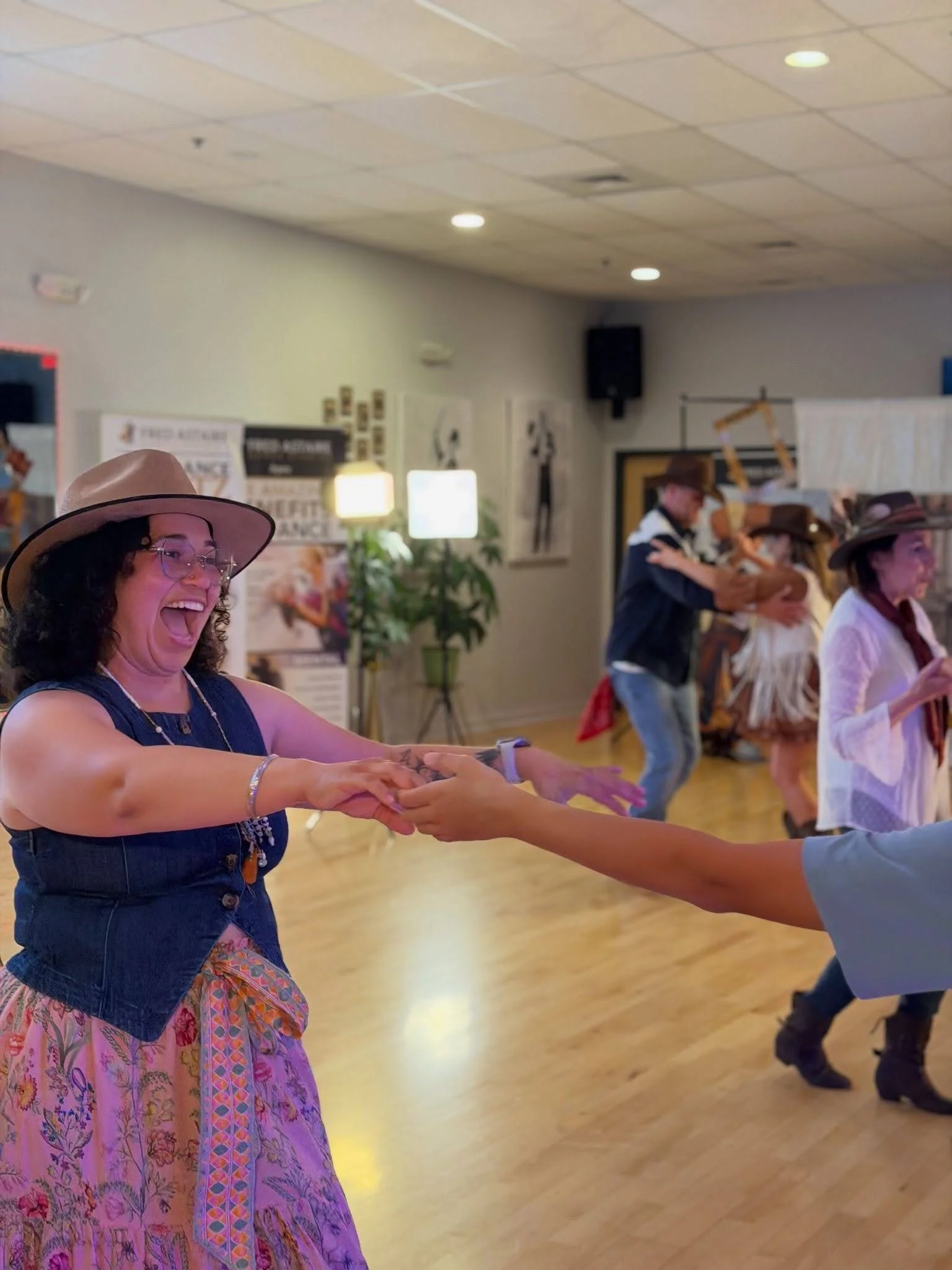 A smiling adult dancer wearing a cowboy hat and colorful outfit enjoys a partner dance in a bright studio while other couples dance in the background, highlighting the fun, welcoming atmosphere of adult dance lessons in Apex.