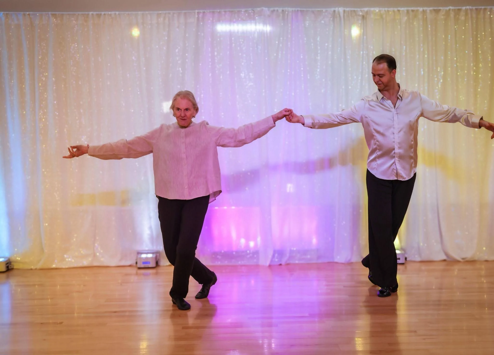 An older adult and an instructor perform a graceful ballroom dance routine on a polished dance floor with arms extended, demonstrating confidence, balance, and the welcoming experience of ballroom dance lessons in Apex.