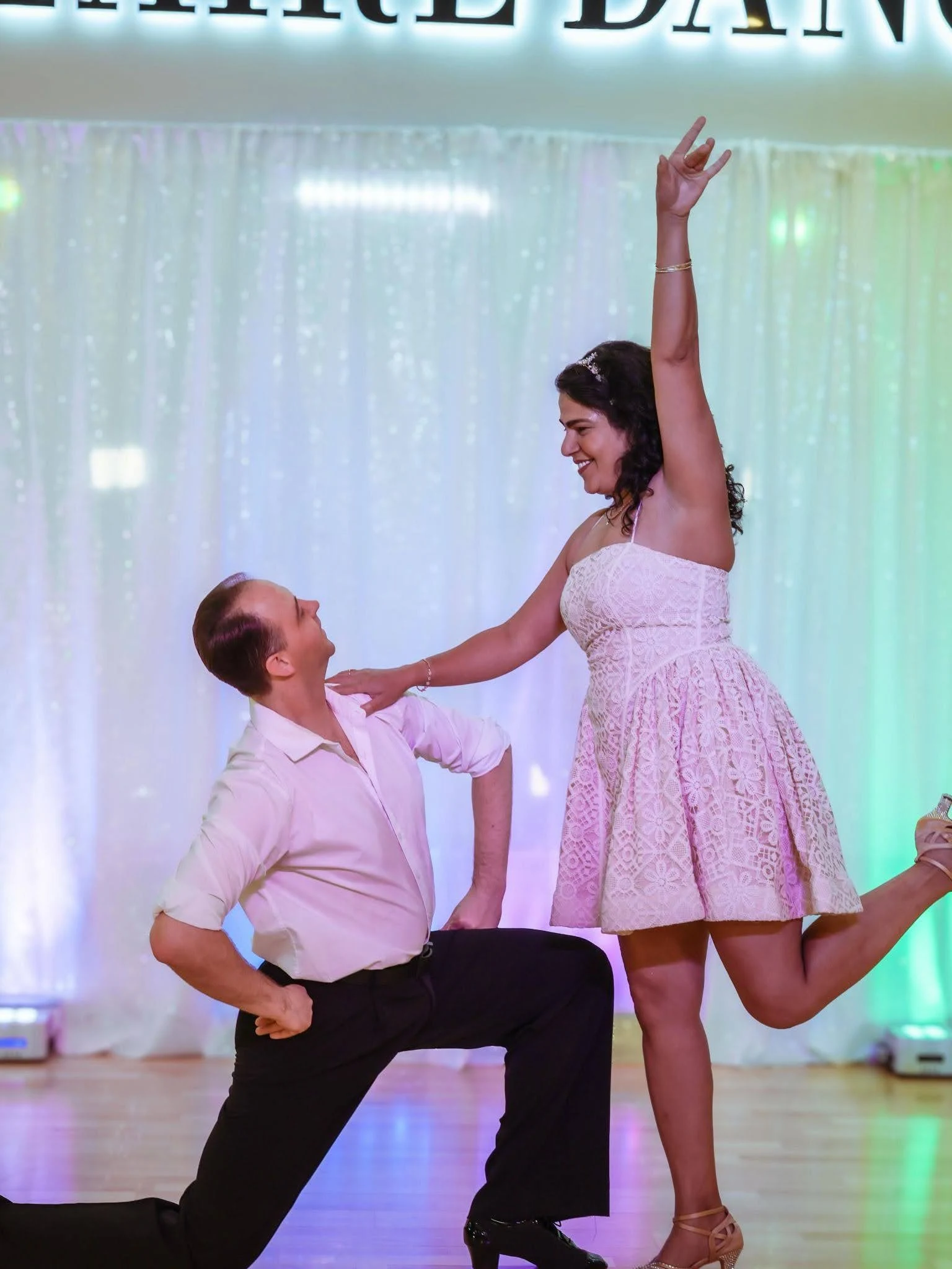 A couple performs a joyful ballroom dance pose on a dance floor, with the woman in a white lace dress raising one arm while smiling at her partner, who kneels and looks up at her. The elegant setting highlights the fun and confidence gained through dance classes in Apex.
