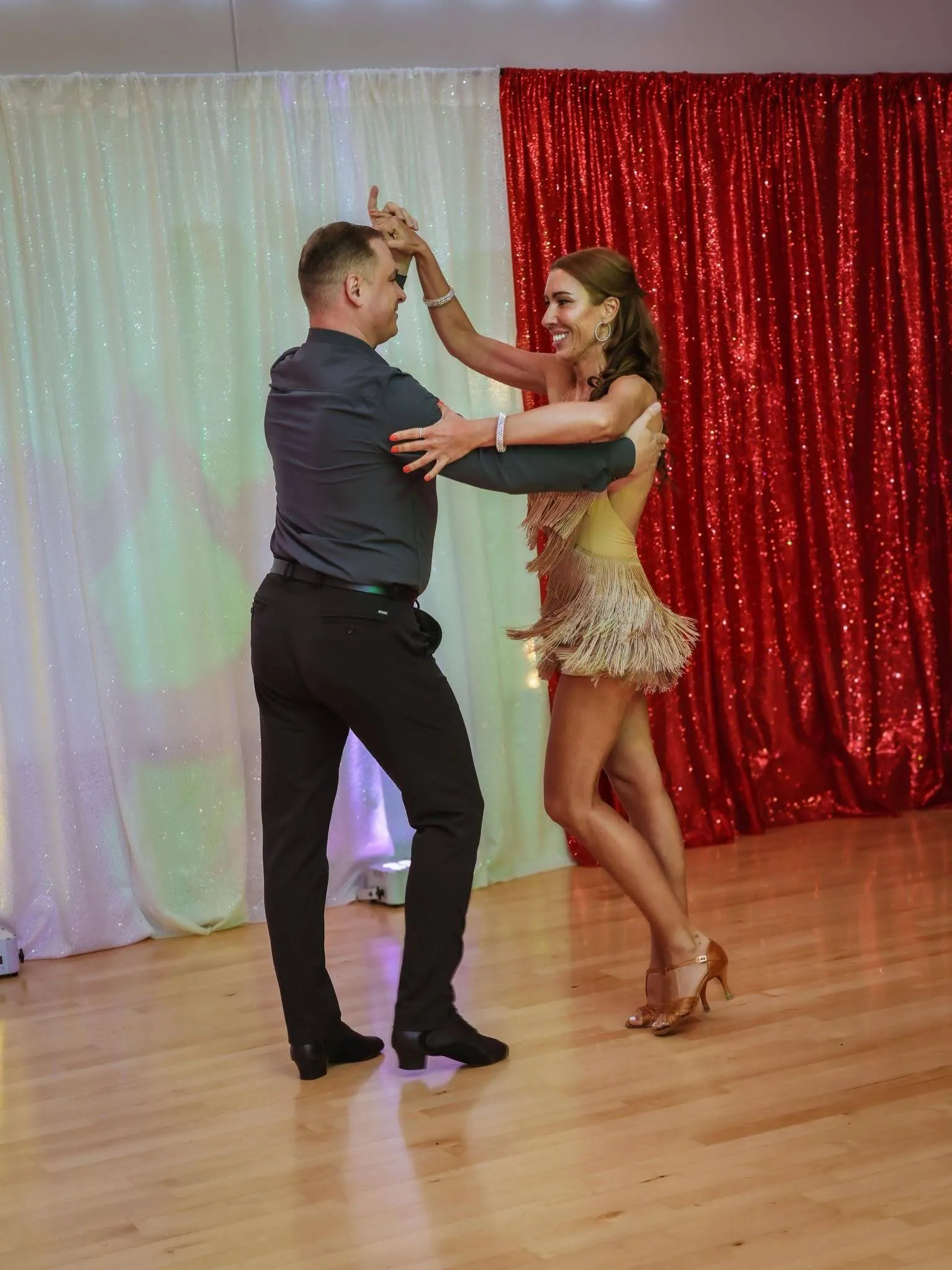 A smiling couple practices a partner dance during adult dance lessons in Apex, with the woman in a gold fringe Latin dress and heels and the man in a dark shirt and trousers performing a graceful turn on a polished dance floor in front of sparkling red and white backdrop curtains.