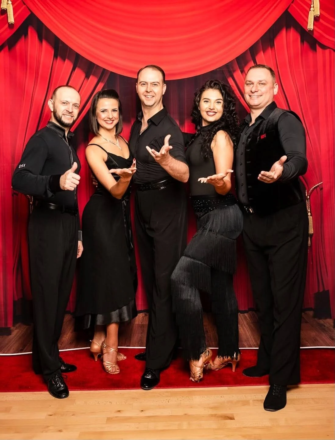 A group of five smiling ballroom and Latin dance instructors pose together in elegant black attire with welcoming gestures in front of a red theater-style curtain, showcasing one of the Best Dance Studios in Apex for Beginners in a friendly and professional atmosphere.