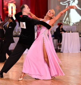 Ballroom Dance Competition: A couple performs an elegant ballroom dance routine, with the woman in a flowing pink gown and the man in a black suit on a competition dance floor.