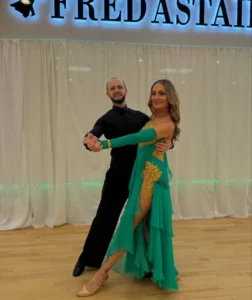 Ballroom Dance Competition: A couple poses in a ballroom dance hold, with the woman wearing an elegant teal and gold gown and the man dressed in black on a wooden dance floor.