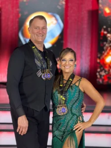 Five Medals on Rhythm Day: A smiling ballroom dance couple proudly displays their five competition medals, celebrating their achievements at Rhythm Day, with the woman wearing a sparkling teal dress and the man dressed in black.