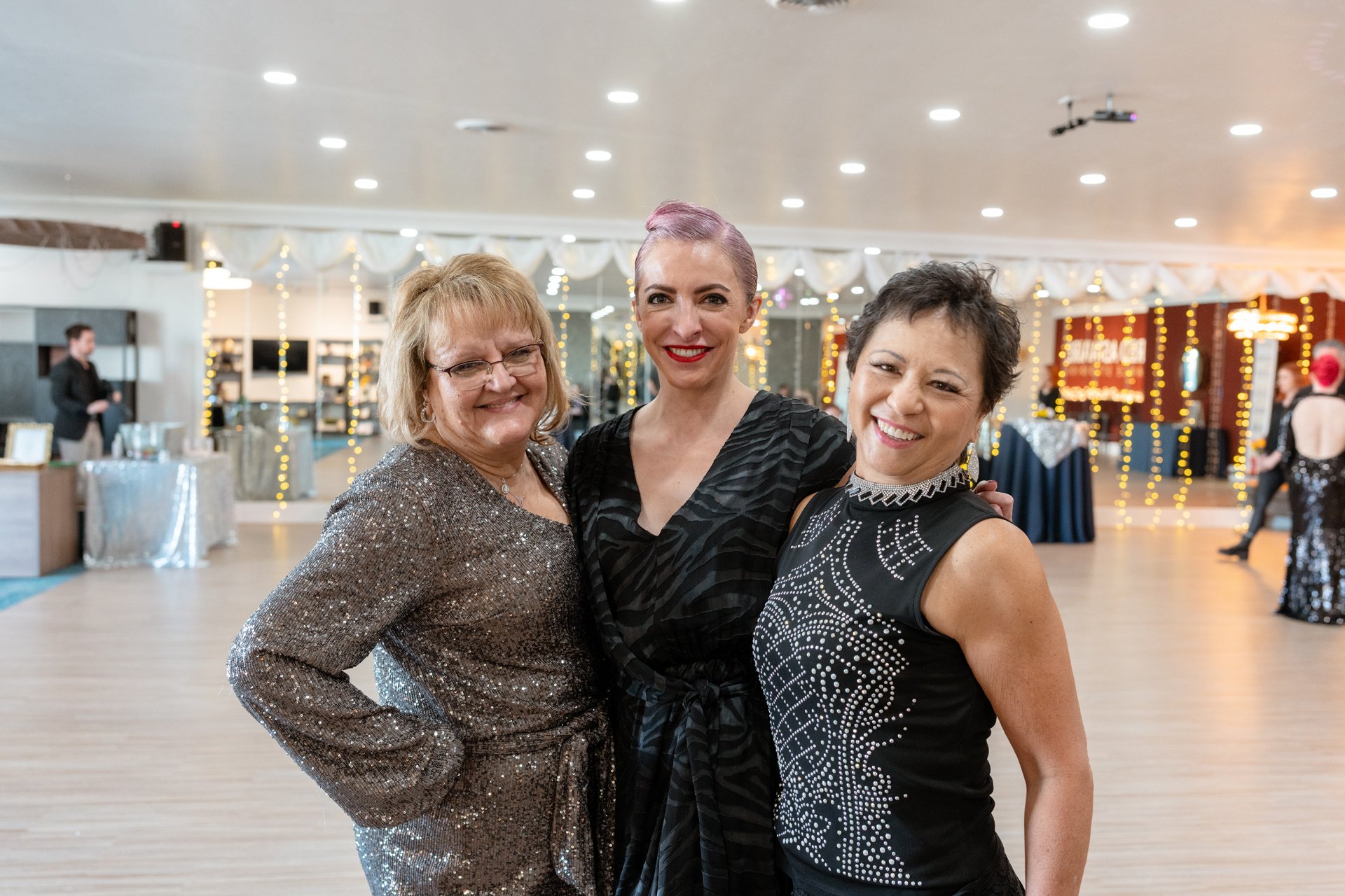 Three friends in sparkly ballroom dance outfits smile on the dance floor at Fred Astaire Boise.