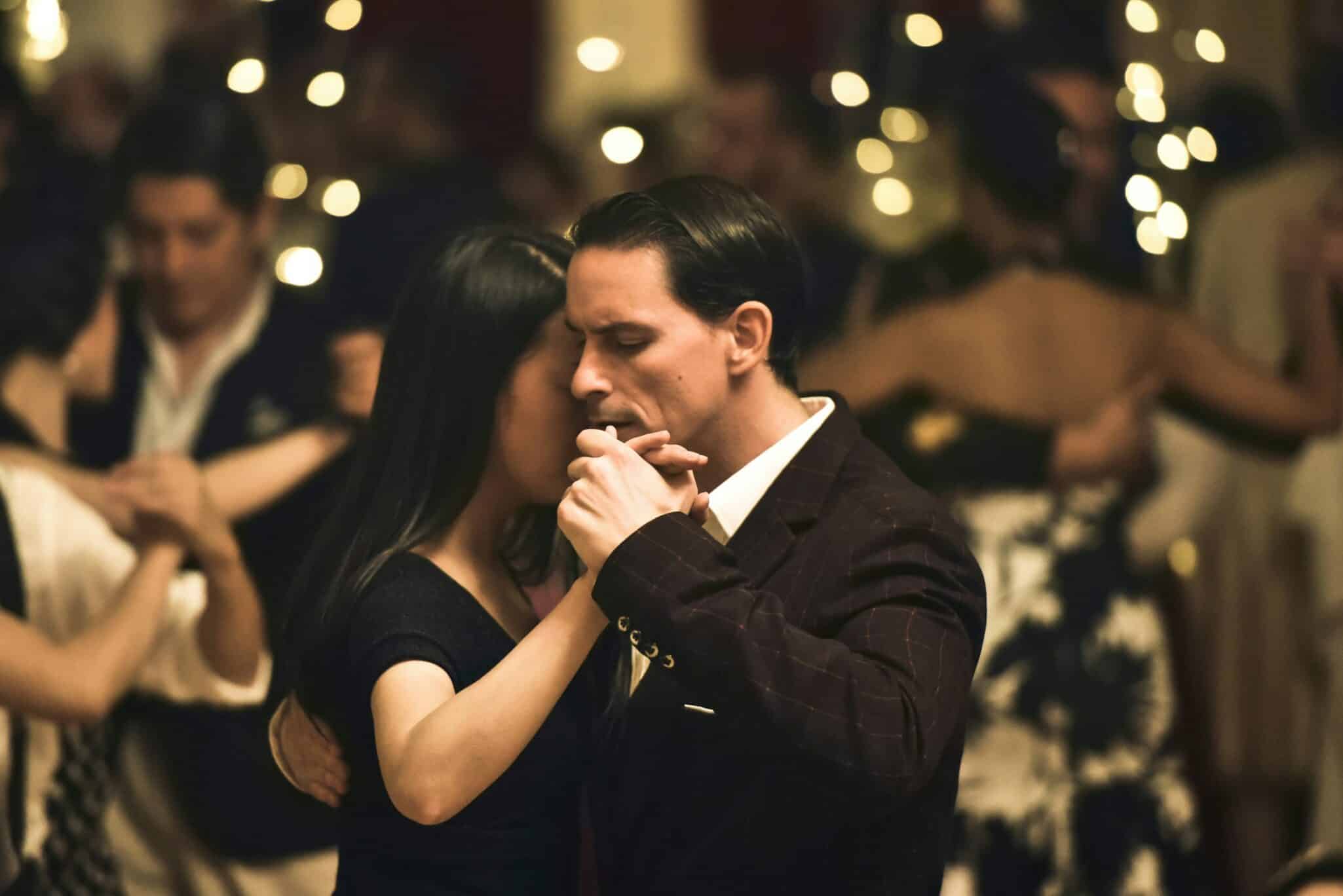 Man and woman practicing ballroom dances in a large room with other dancers.