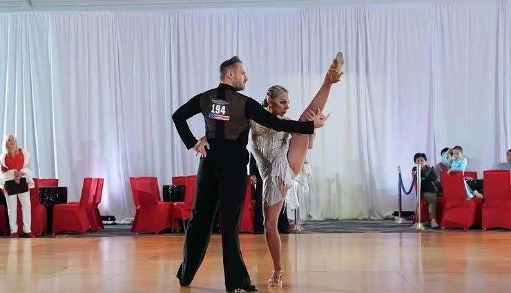 Professional dancers perform a dramatic Pro-Am Ballroom Dancing in Washington routine during a ballroom competition, with the female dancer executing a high leg extension while her partner poses beside her on a polished dance floor in front of an audience.