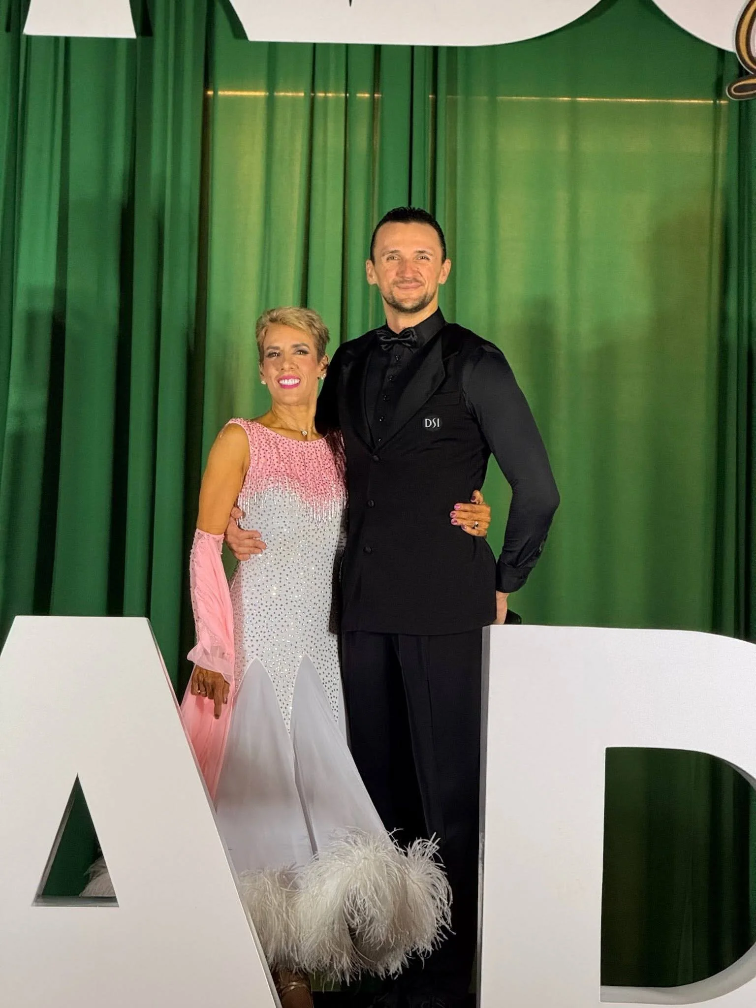 A ballroom dance couple posing in formal competition attire on stage during a dance event, showcasing the skills learned through dance classes in Orlando FL.