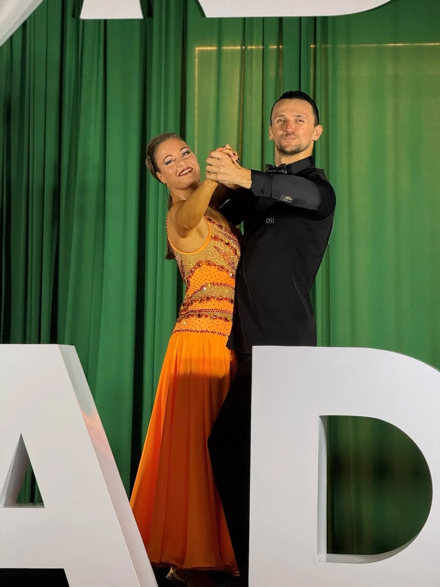 A ballroom dance couple performing together in front of a green curtain, featuring a woman in an orange dress and a man in black attire during Ballroom Dancing in Orlando.