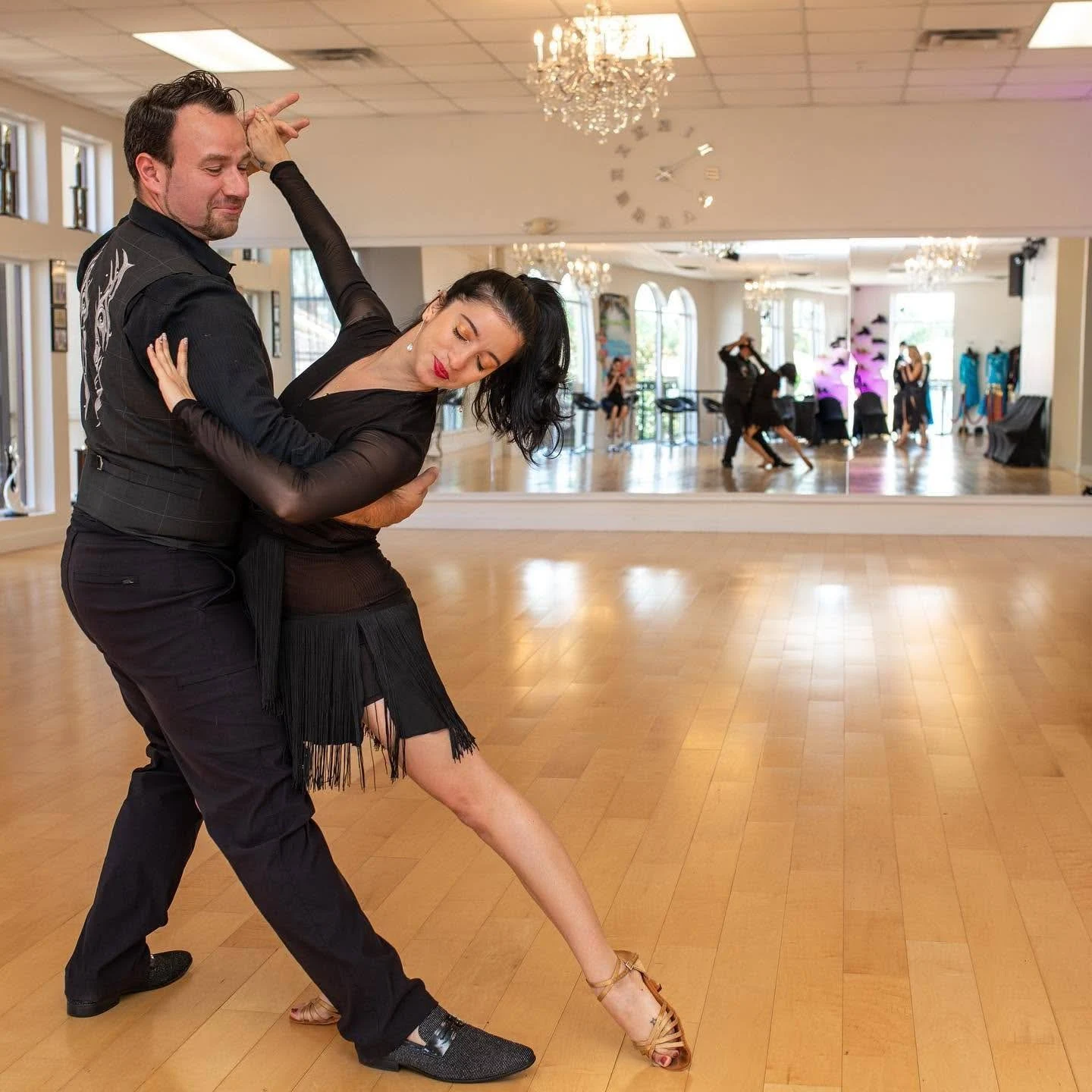 Date Night Idea in Orlando, a couple in black dancing a Latin routine on a wood floor in a bright dance studio with a chandelier