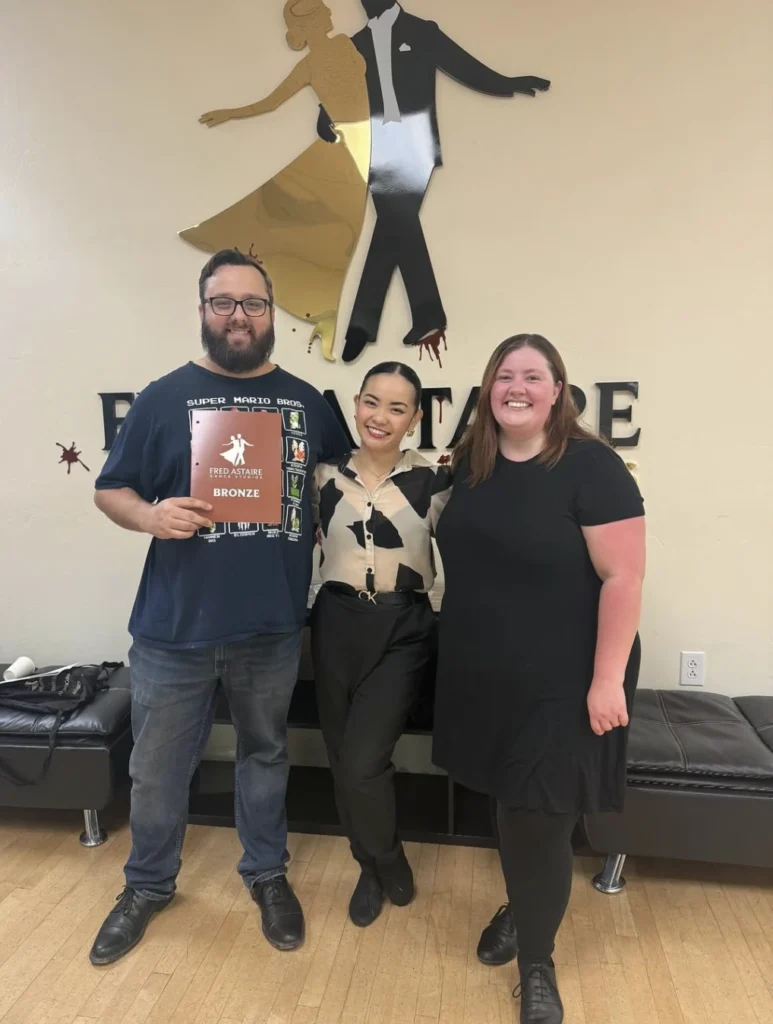 Three individuals standing in a dance studio, one holding a “Fred Astaire Bronze” plaque, celebrating achievement in dance lessons.