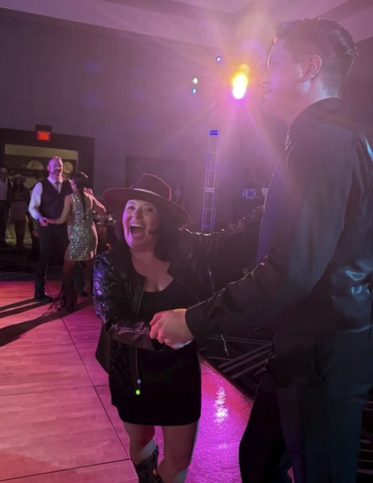 A joyful couple dancing under vibrant purple and yellow lights at a lively indoor event, surrounded by others enjoying the celebration—capturing the energy and connection of dancing.