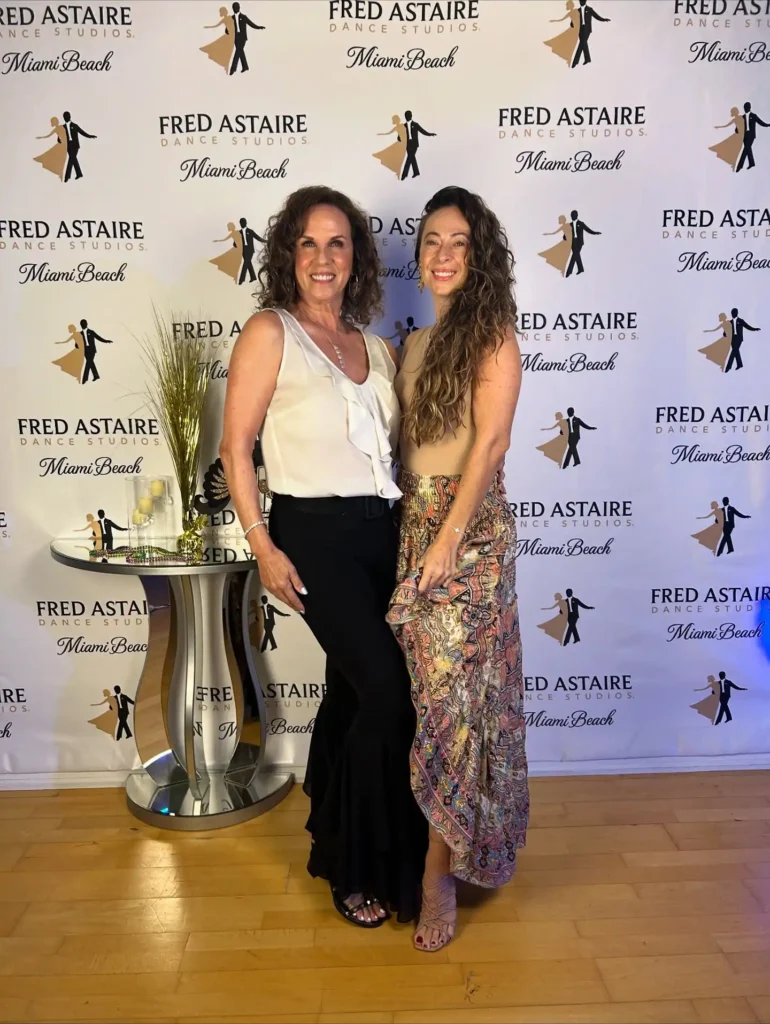 Two women standing together and smiling at a Fred Astaire Dance Studio Miami Beach event, posing in front of a branded backdrop that promotes latin dance lessons.
