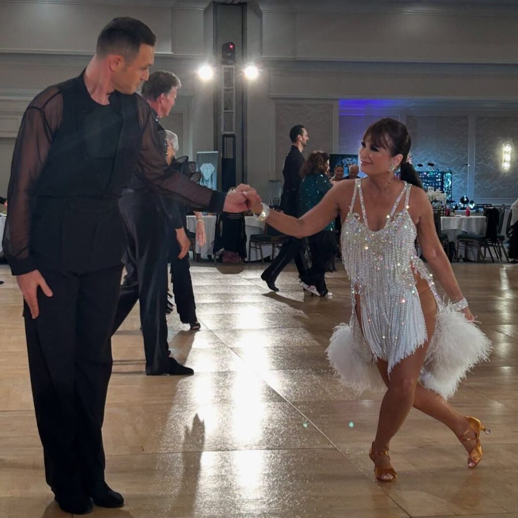 A male and female dancer performing paso doble at a ballroom event. The man wears a black outfit with sheer sleeves; the woman wears a white fringe dress with feathers and tan dance heels. They are mid-motion on a well-lit dance floor surrounded by tables and other dancers.
