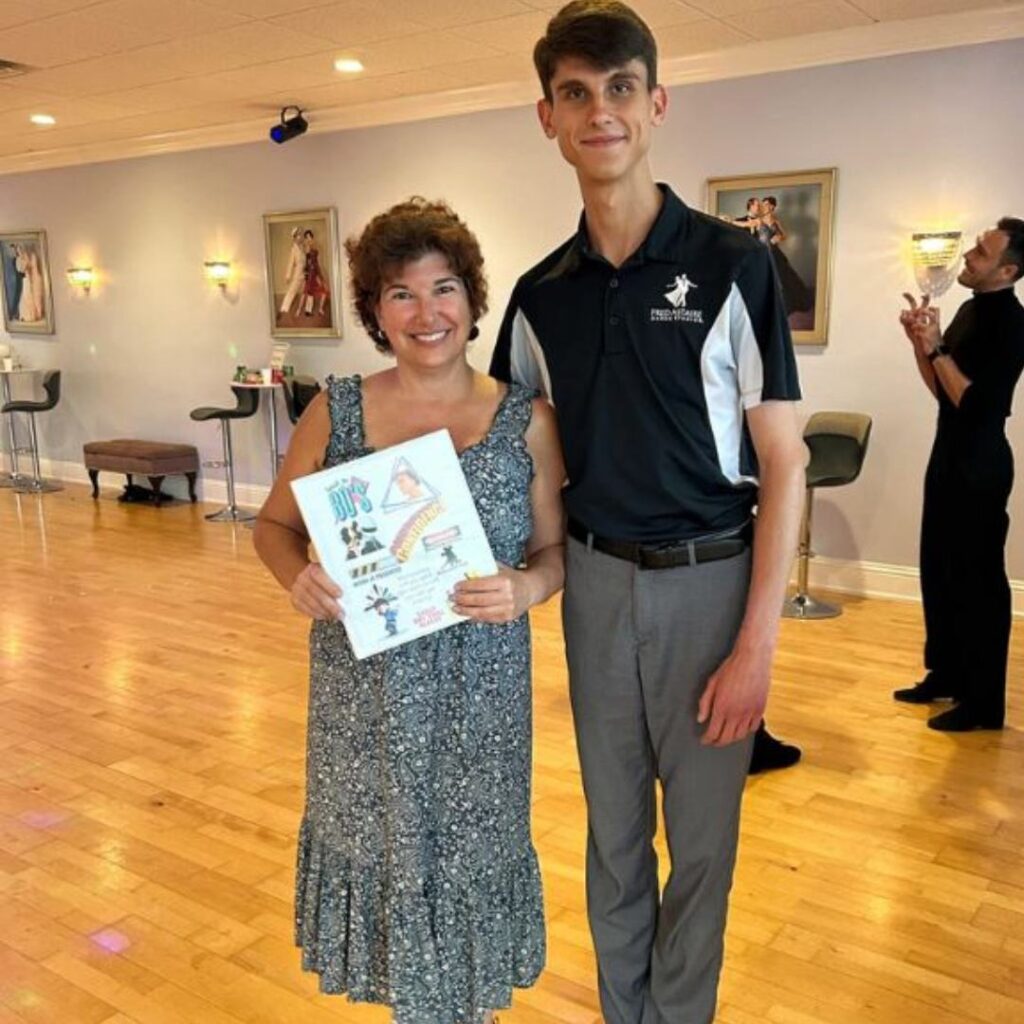 Two people standing in a dance studio during a celebration related to waltz dance classes. The woman holds a decorated certificate; the man wears a Fred Astaire Dance Studios polo shirt.