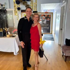 Dance Lessons for Adults: A smiling adult couple poses together at a dance studio, with the woman wearing a red fringe dress and the man dressed in black, celebrating their ballroom dancing journey.