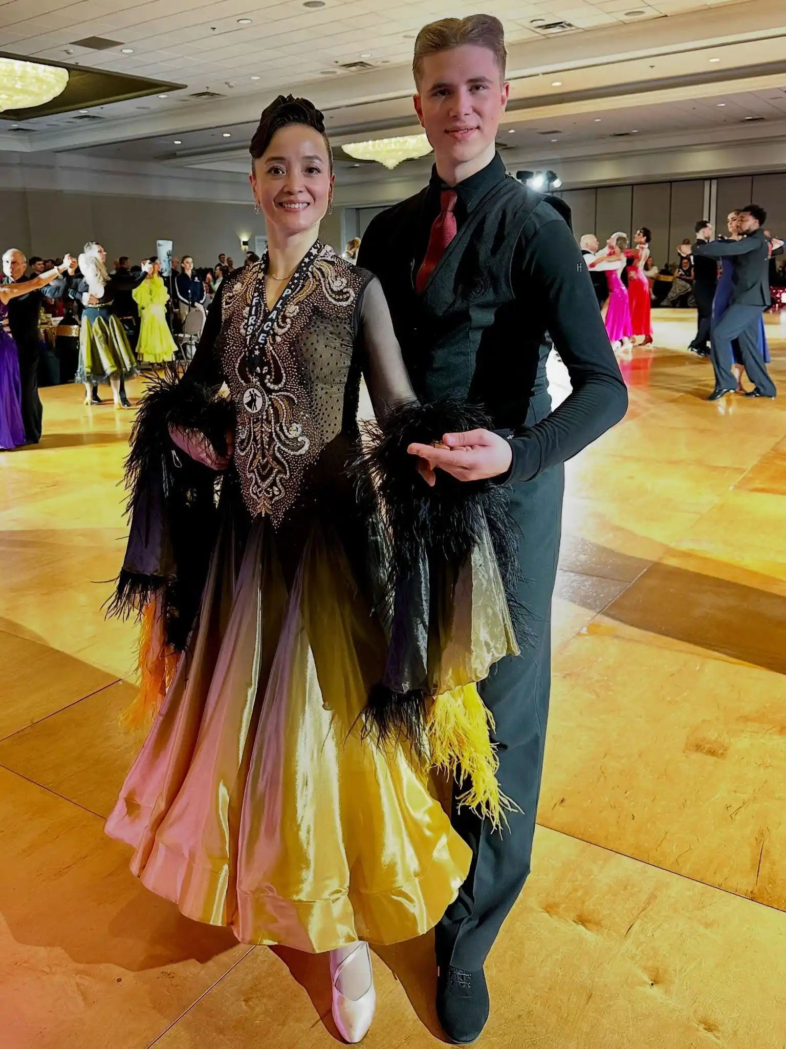 Pro-Am Dancing in Morrisville featuring a ballroom dance couple posing on the competition floor, with the female dancer wearing a black embellished gown and the male partner dressed in a black outfit with a red tie.