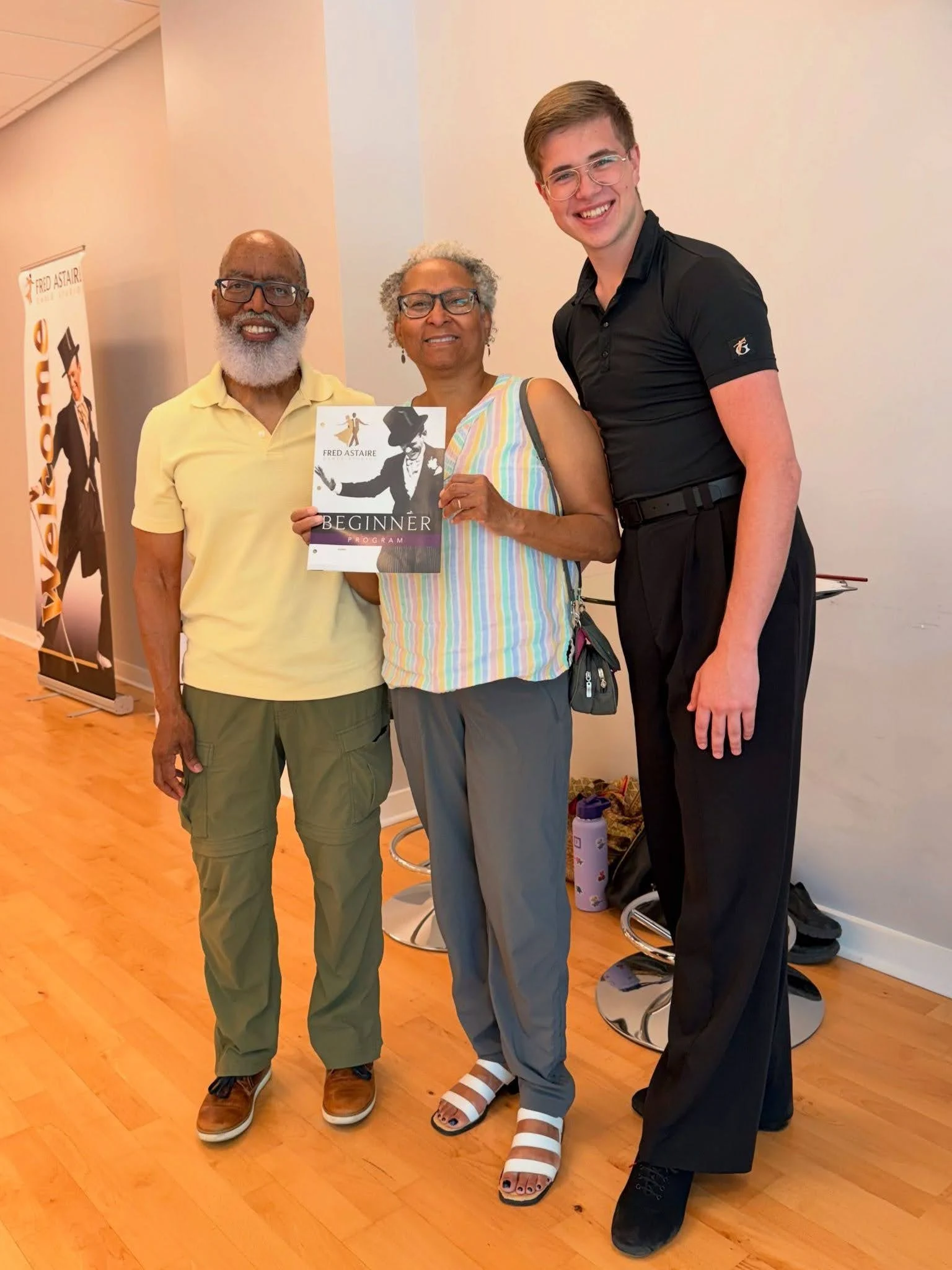 Start Dancing as an Adult in Morrisville: Two new adult dance students smile alongside a ballroom instructor while holding a beginner program guide at Fred Astaire Dance Studios, celebrating the start of their dance journey.