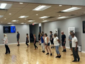 Adults participating in a group dance class at Fred Astaire Dance Studios, practicing dance steps together in a spacious studio with a wooden floor.