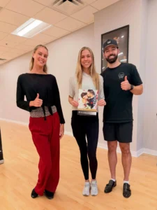 Three adults smiling in a dance studio, holding a dance-related booklet, illustrating the Difference Between Ballroom and Latin Dancing.