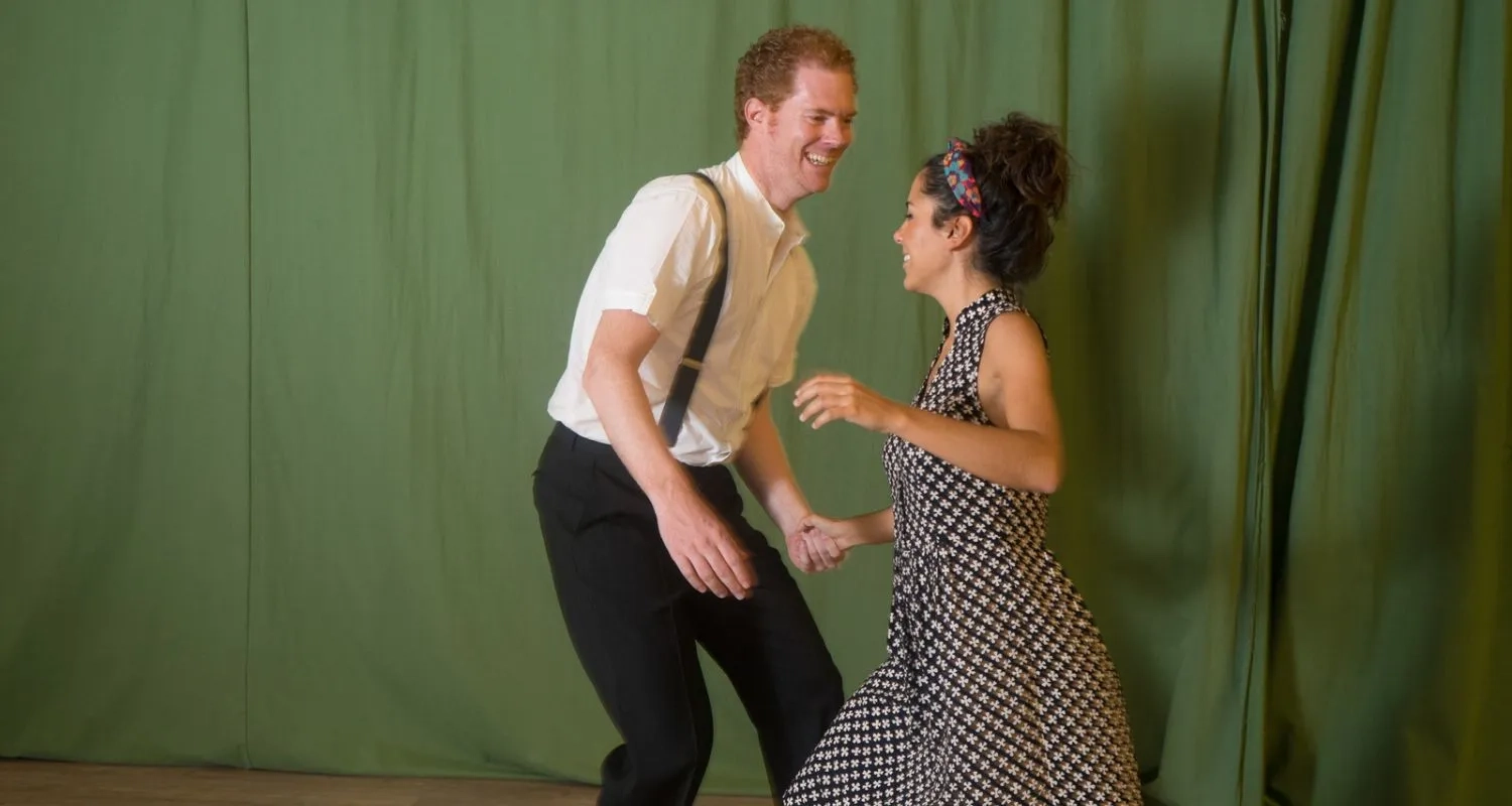 A man and woman in modern dance attire practice an East Coast Swing routine within a well-lit studio.