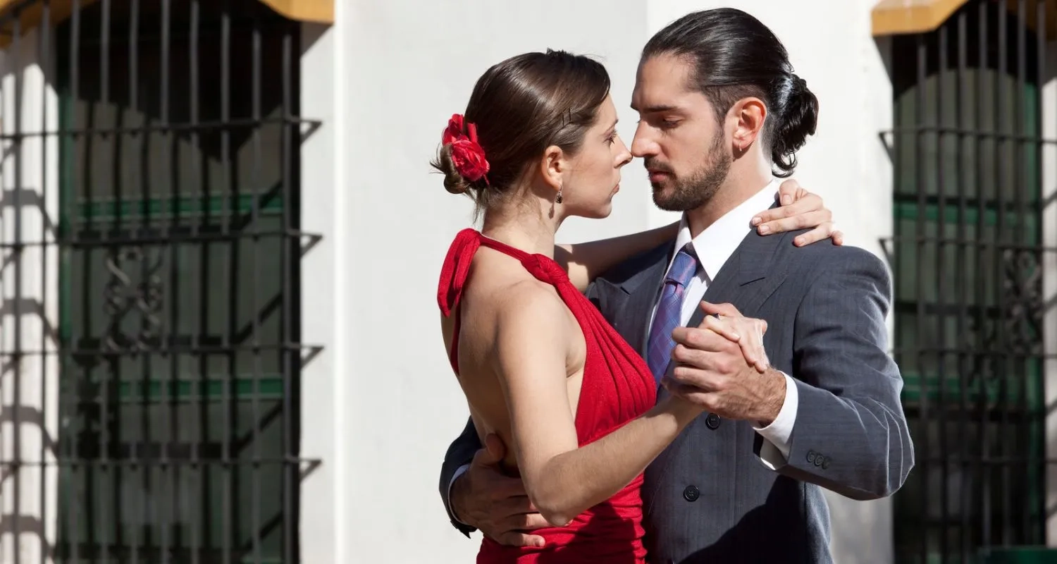 Couple dancing Argentine Tango in a close embrace on a sunlit street in front of a green iron gate