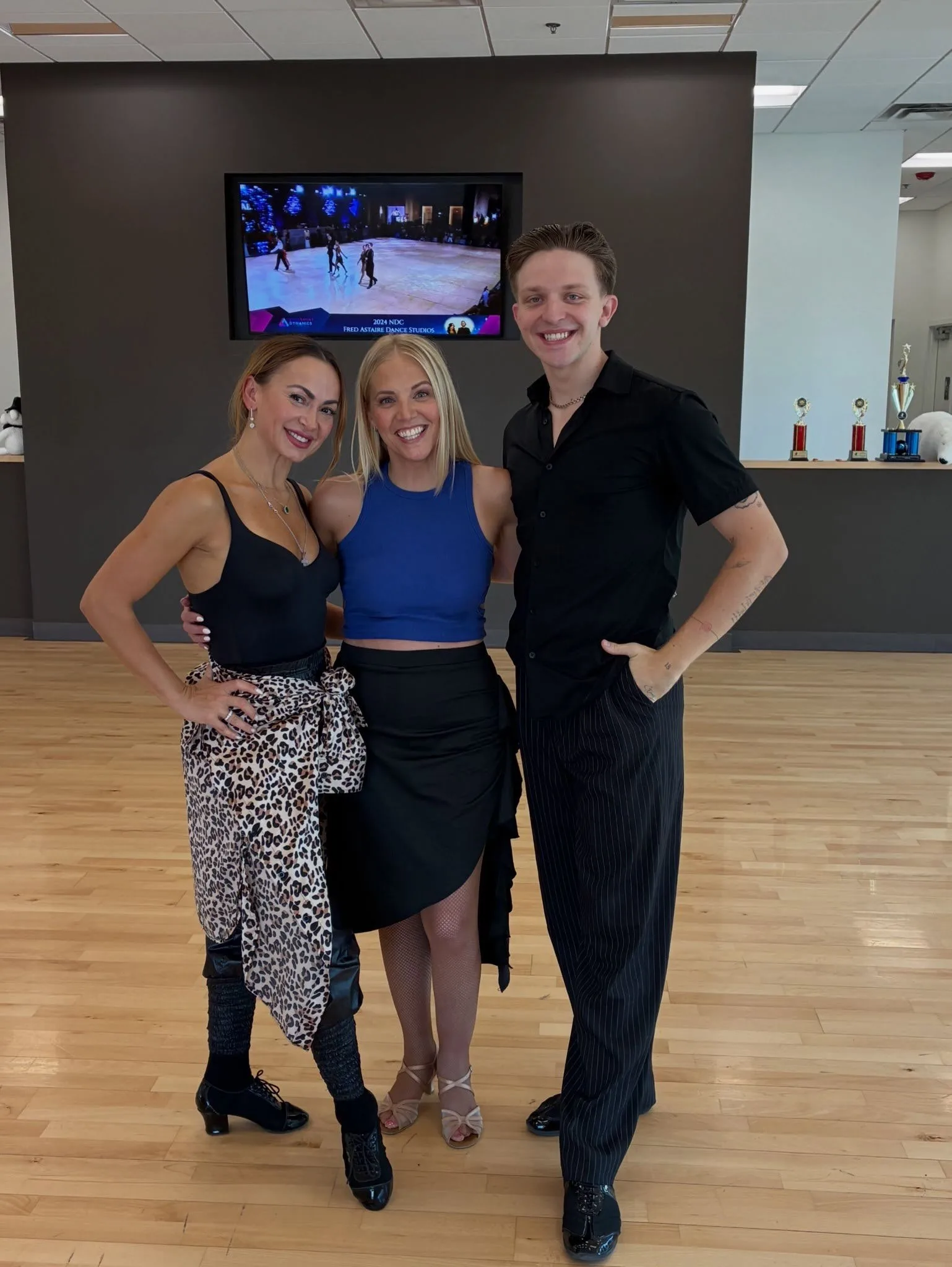 Three smiling dance instructors pose on the wooden floor of a bright ballroom studio offering dance lessons for couples, with competition trophies and a ballroom video screen behind them.