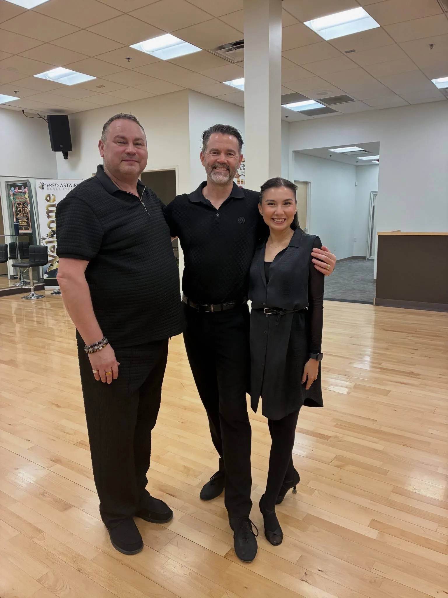 Three smiling dance instructors dressed in black pose on the hardwood floor of a Fred Astaire Dance Studios ballroom, where students can learn rumba in Peoria.