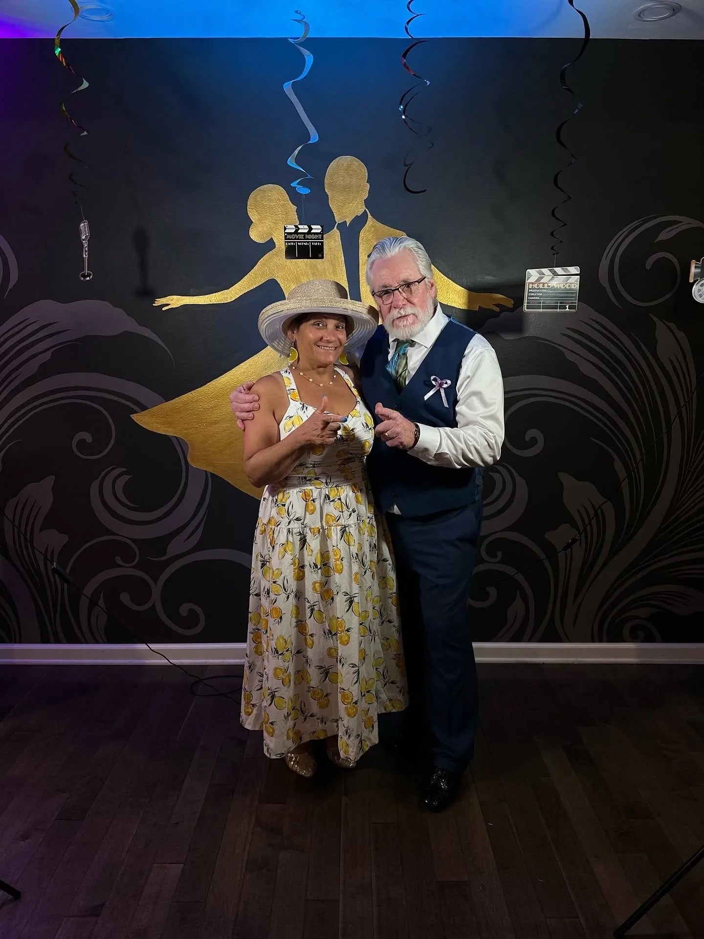 A smiling couple poses together in front of a Hollywood-themed backdrop featuring a gold dancing silhouette. The woman wears a white floral dress with a wide-brimmed hat, while the man wears a blue suit vest, tie, and white shirt. Both point playfully toward the camera, showcasing the fun and welcoming atmosphere of Beginner Dance Lessons in Saratoga Springs.