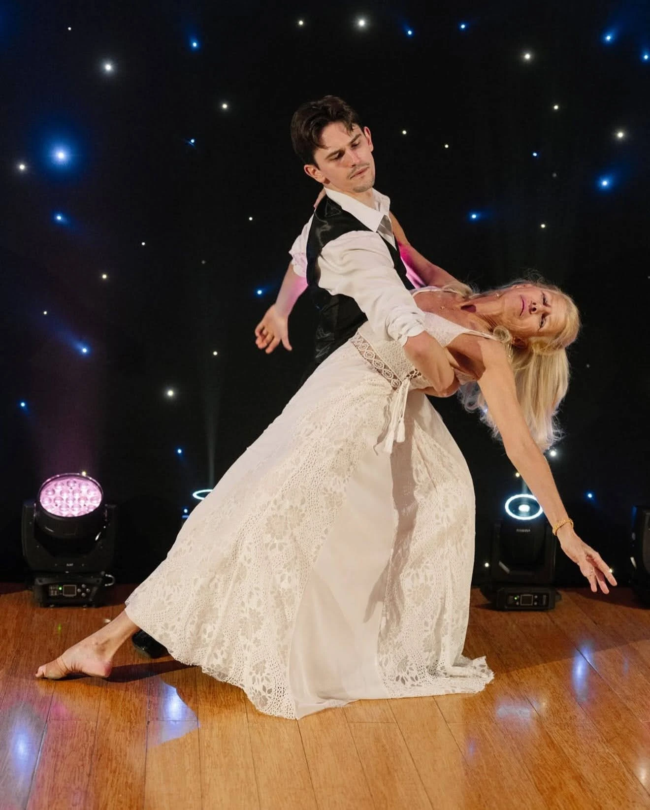 A dance couple performs an elegant ballroom dip on a polished wooden floor, with the lead supporting their partner in a flowing white lace dress against a star-lit backdrop. This graceful performance showcases the style and confidence you can build through Beginner Ballroom Dance Lessons in NYC.