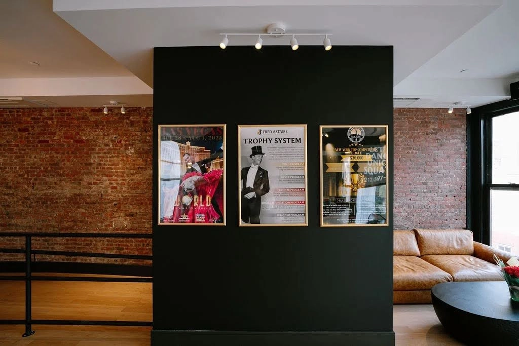 A stylish dance studio lobby features exposed brick walls, framed dance competition posters, modern lighting, and a comfortable seating area, creating a welcoming space for students ready to Learn Salsa in New York City.