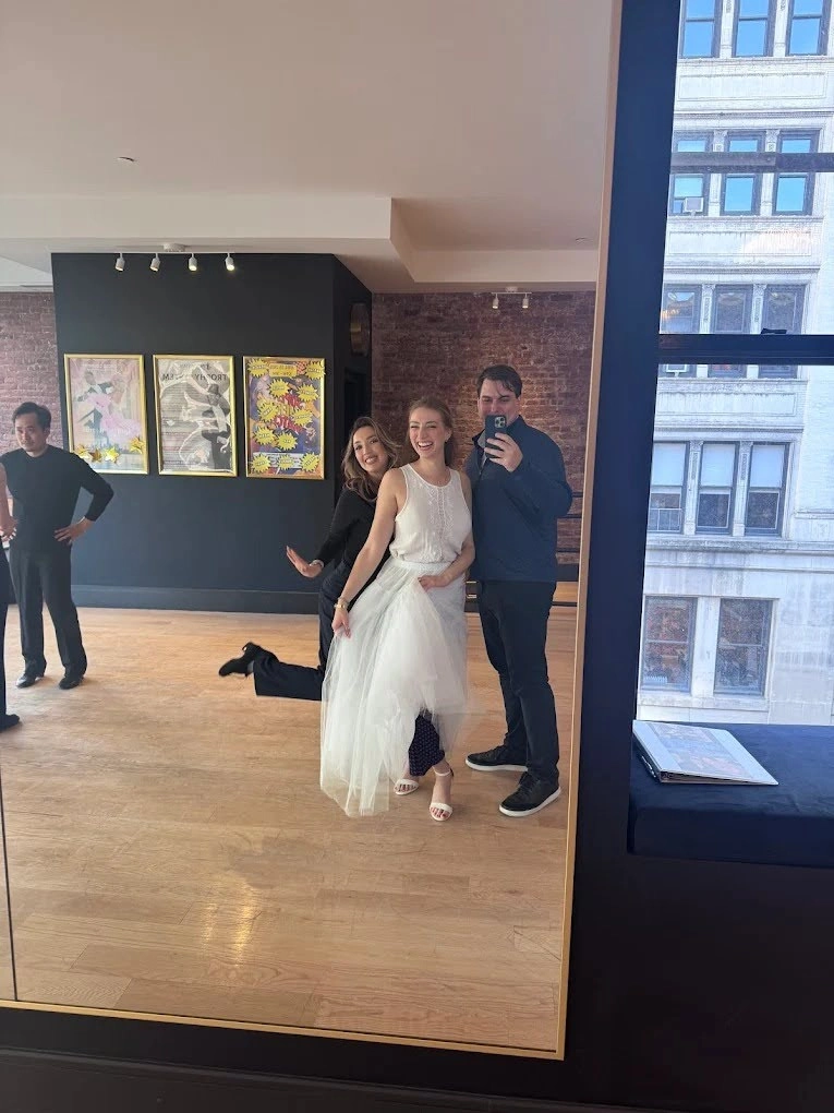 Three people smile while posing for a mirror selfie inside Fred Astaire Dance Studios. One person wears a flowing white dance dress, another playfully lifts a leg behind them, and a third takes the photo with a phone. The spacious studio features hardwood floors, exposed brick walls, framed artwork, and large windows overlooking city buildings.
